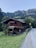 A rustic wooden cabin is nestled in a lush green landscape with mountainous scenery in the background. The cabin, with its dark wooden exterior and visible balconies, is surrounded by green trees and grass. A narrow road runs alongside the cabin, leading toward the mountains.