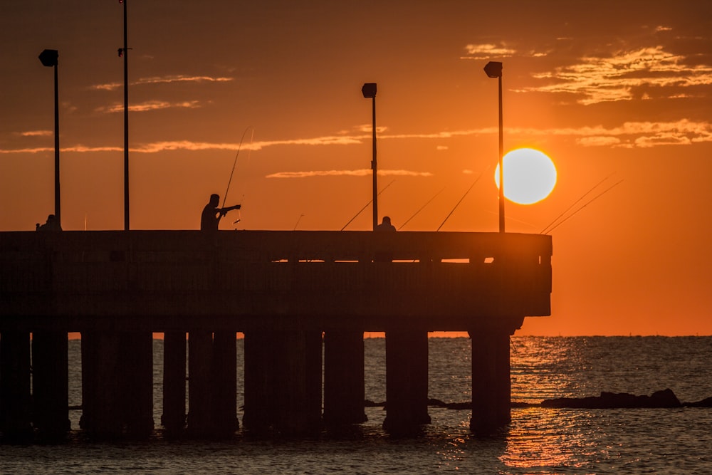 a couple of people fishing on a pier at sunset