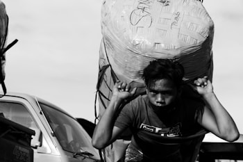 A person is carrying a large, heavily wrapped parcel on their back, using both hands to support it. The scene is captured in black and white, with a vehicle visible in the background, indicating an outdoor setting.