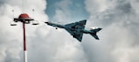 A military jet flies prominently through a cloudy sky, appearing to perform a maneuver near a red and white tower structure. The aircraft has distinctive insignia and markings on its wings and fuselage.