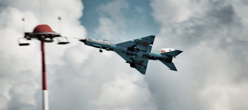 A military jet flies prominently through a cloudy sky, appearing to perform a maneuver near a red and white tower structure. The aircraft has distinctive insignia and markings on its wings and fuselage.