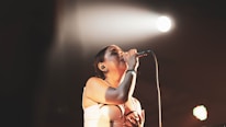 A person is singing passionately into a microphone under a spotlight with a dark background. The intensity of their expression suggests they are deeply engaged with the performance.