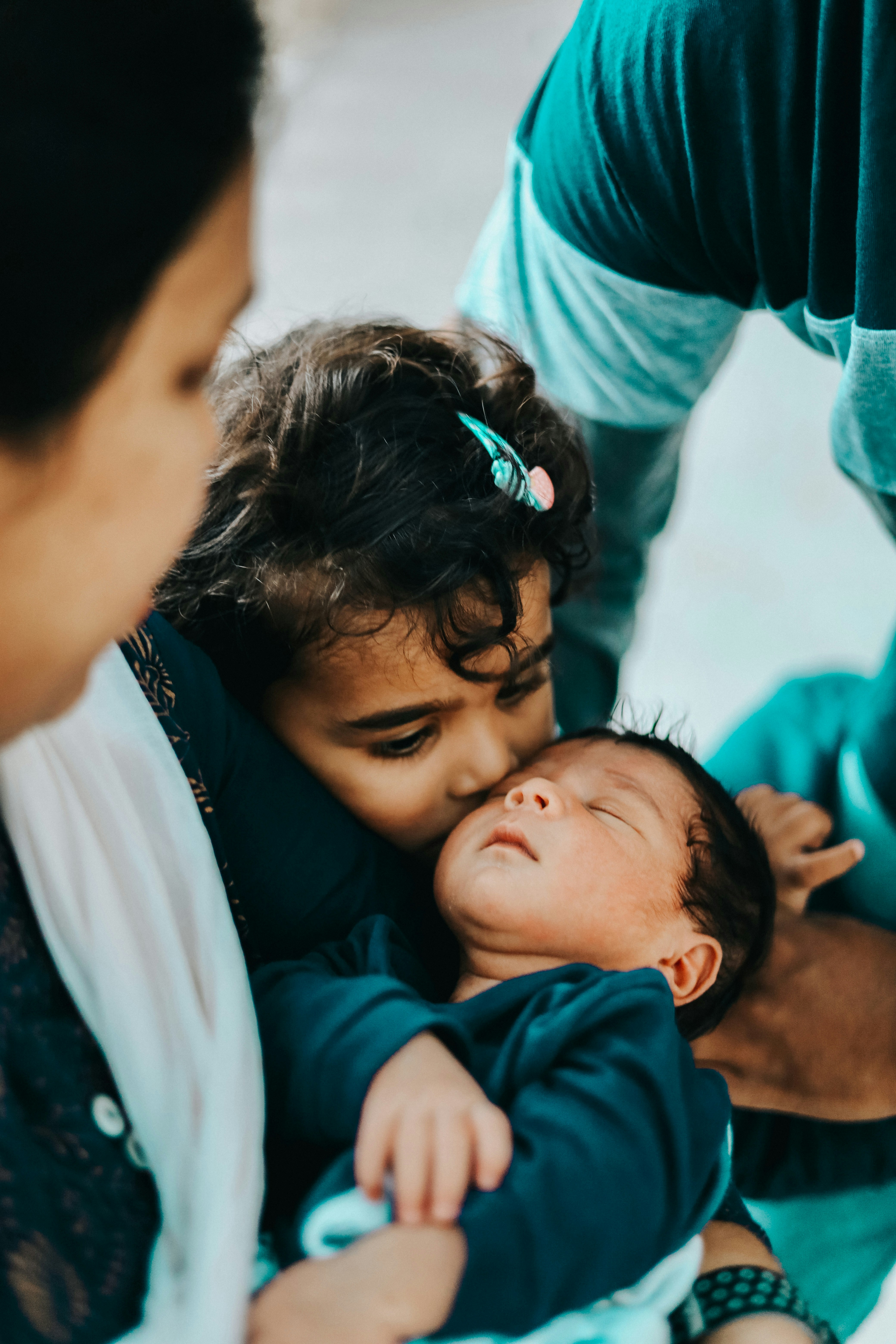 A young girl with curly hair and a colorful hair clip gently kisses her newborn sibling, who is peacefully sleeping in someone's arms. Family members surround them