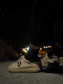 A pair of shoes with a distinct logo are worn by someone standing on a concrete surface during nighttime. The background is slightly blurred with glowing city lights in the distance, creating a contrast between the dark surroundings and the illuminated footwear.