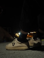 A pair of shoes with a distinct logo are worn by someone standing on a concrete surface during nighttime. The background is slightly blurred with glowing city lights in the distance, creating a contrast between the dark surroundings and the illuminated footwear.