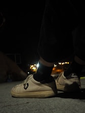 A pair of shoes with a distinct logo are worn by someone standing on a concrete surface during nighttime. The background is slightly blurred with glowing city lights in the distance, creating a contrast between the dark surroundings and the illuminated footwear.
