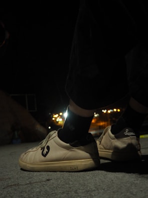 A pair of shoes with a distinct logo are worn by someone standing on a concrete surface during nighttime. The background is slightly blurred with glowing city lights in the distance, creating a contrast between the dark surroundings and the illuminated footwear.