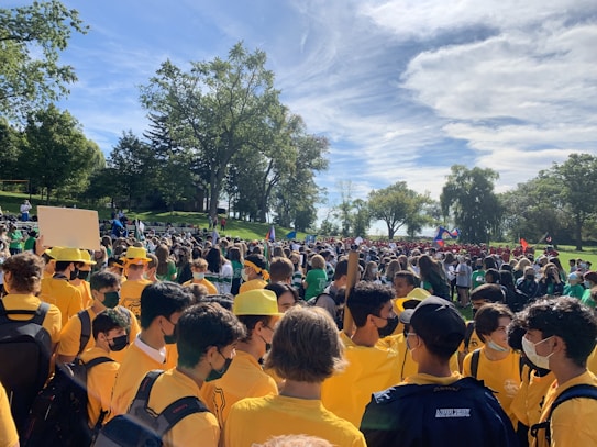 A large group of people gathered outdoors in a park-like setting, many wearing matching yellow shirts and hats. Trees and a clear sky are visible in the background, along with other groups of people wearing different colored shirts, suggesting a coordinated event or gathering.