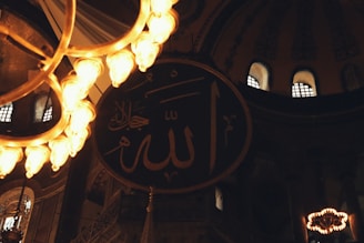 A dimly lit interior with a large circular calligraphy of Arabic script hanging on the wall. The script is surrounded by an ornate chandelier with multiple glowing lights that illuminate the foreground. In the background, there are arched windows streaming soft light into the space.
