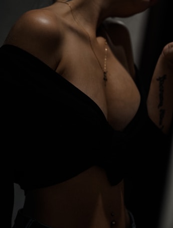 A close-up of a person's upper body with an off-the-shoulder black top, revealing a tattoo on the arm and a necklace. The body is toned and there is a belly piercing.