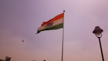 A large Indian national flag is hoisted on a pole against a clear sky. A bird is flying near the flag. There is also a street lamp on the right side of the image.