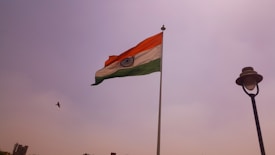 A large Indian national flag is hoisted on a pole against a clear sky. A bird is flying near the flag. There is also a street lamp on the right side of the image.