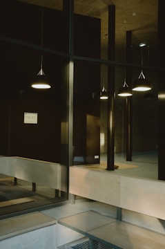 A modern interior space featuring a series of hanging pendant lights casting a soft glow. The area includes large glass panels and a combination of wood and metal finishes. A minimalistic design is evident, with clean lines and neutral tones.