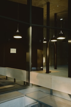 A modern interior space featuring a series of hanging pendant lights casting a soft glow. The area includes large glass panels and a combination of wood and metal finishes. A minimalistic design is evident, with clean lines and neutral tones.
