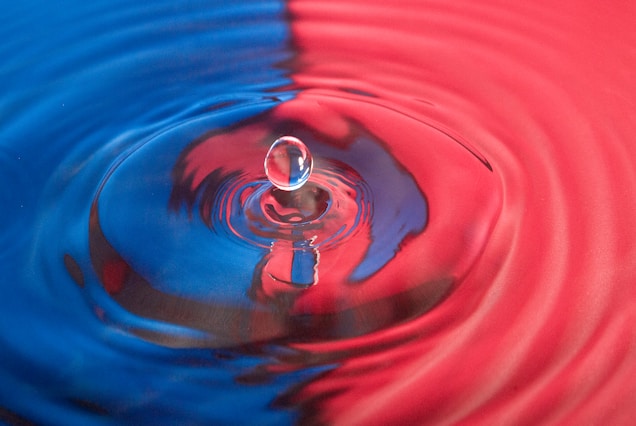 A droplet of water is caught in motion as it impacts a surface, creating ripples that spread outwards. The surface is divided into two contrasting colors, blue and red, with the droplet casting a reflection at the center. The image captures the fluid dynamics and symmetry of the splash.