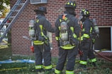 Three firefighters in full gear stand in front of a brick building. They wear helmets and oxygen tanks, and appear to be in the process of assessing a situation or preparing for action. A ladder is propped against the building and a hose lies on the ground near them.