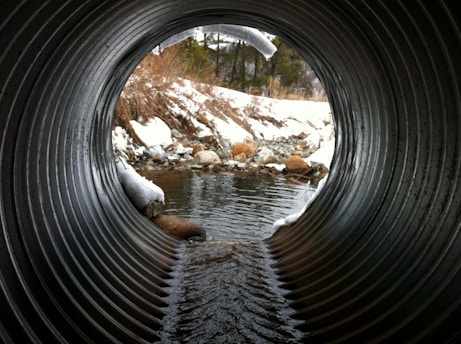 A view from inside a culvert pipe looking out onto a snowy landscape. The pipe is metallic and ridged, with some snow and ice around its edges. Beyond the pipe, a small stream flows over rocks, flanked by snow-covered banks and sparse vegetation. In the distance, evergreen trees rise against a cloudy sky.