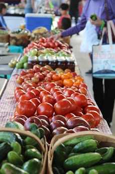 A vibrant display of fresh produce including tomatoes and cucumbers is arranged on a market table. A person is selecting vegetables, and jars of preserved goods are in the background. Shoppers and vendors move about in the distance.