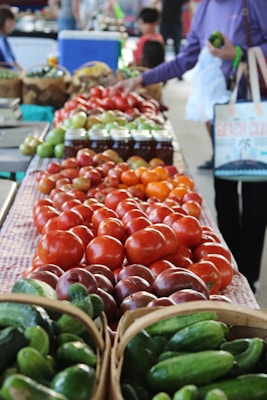 A vibrant display of fresh produce including tomatoes and cucumbers is arranged on a market table. A person is selecting vegetables, and jars of preserved goods are in the background. Shoppers and vendors move about in the distance.
