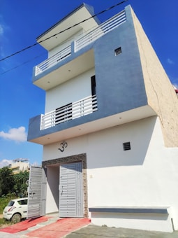 A multi-story modern house with white and gray exterior colors. There is a large gate leading to an entrance, with a car parked on the left side. The house has balconies on the upper floors with white railings and a symbol 'Om' above the main entrance.