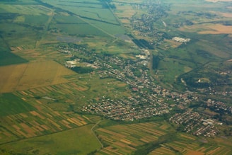 An aerial view of a rural landscape with patchwork fields and a small town. The fields are arranged in various geometric shapes and colors, surrounded by greenery. A network of roads and buildings form the town, merging into the countryside.