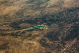 An aerial view of a vast landscape featuring a winding body of water surrounded by hilly or mountainous terrain. The landscape includes textured surfaces indicating variations in vegetation and rock formations, with a small town or settlement visible at one edge.
