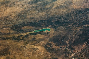 An aerial view of a vast landscape featuring a winding body of water surrounded by hilly or mountainous terrain. The landscape includes textured surfaces indicating variations in vegetation and rock formations, with a small town or settlement visible at one edge.