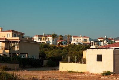 A serene residential area with multiple Mediterranean-style villas featuring terracotta roof tiles and pastel-colored walls. The houses are surrounded by lush greenery and are situated under a clear blue sky with hills in the background.