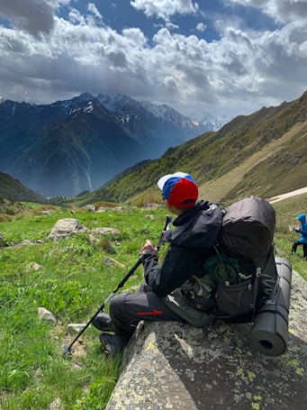 A person wearing outdoor gear and a colorful cap sits on a rock in a lush, mountainous landscape. They carry a large backpack, hiking poles, and a rolled-up sleeping mat, suggesting they are hiking or camping. Snow-capped mountains rise in the background under a partially cloudy sky, creating a serene and adventurous atmosphere.