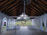 An outdoor covered structure with a high, open ceiling and wooden beams, featuring sheer white drapes along the sides. The flooring is wooden, and there are several rows of white folding chairs positioned facing outward toward a lush, green vineyard outside. A circular metal chandelier hangs from the center of the ceiling.