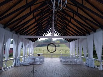 An outdoor covered structure with a high, open ceiling and wooden beams, featuring sheer white drapes along the sides. The flooring is wooden, and there are several rows of white folding chairs positioned facing outward toward a lush, green vineyard outside. A circular metal chandelier hangs from the center of the ceiling.