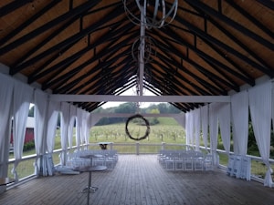 An outdoor covered structure with a high, open ceiling and wooden beams, featuring sheer white drapes along the sides. The flooring is wooden, and there are several rows of white folding chairs positioned facing outward toward a lush, green vineyard outside. A circular metal chandelier hangs from the center of the ceiling.