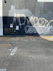 A concrete wall with multiple graffiti tags in various styles and colors, primarily in shades of white, yellow, and black. The wall appears to have textured paint streaks running down from the top. In front of the wall is an empty parking lot with visible parking lines and faded writing on the ground.
