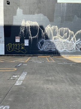 A concrete wall with multiple graffiti tags in various styles and colors, primarily in shades of white, yellow, and black. The wall appears to have textured paint streaks running down from the top. In front of the wall is an empty parking lot with visible parking lines and faded writing on the ground.