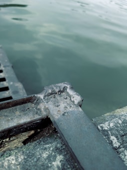A close-up of a metal structure with rust and wear, positioned near the water's edge. The water appears calm with subtle ripples, and the metal shows signs of oxidation and corrosion. Part of the structure seems to be a grate or railing.