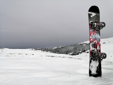 A snowboard with colorful geometric patterns stands upright in a snowy landscape. The background features snow-covered hills and a cloudy sky, creating a serene winter scene.
