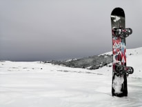 A snowboard with colorful geometric patterns stands upright in a snowy landscape. The background features snow-covered hills and a cloudy sky, creating a serene winter scene.
