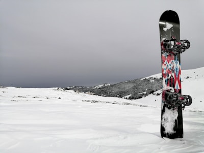 A snowboard with colorful geometric patterns stands upright in a snowy landscape. The background features snow-covered hills and a cloudy sky, creating a serene winter scene.