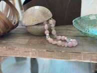A rustic wooden table hosts an assortment of decorative items. A round stone object with a rough texture is positioned next to a pink rose quartz beaded necklace. On the right, a light green glass bowl with intricate patterns sits, while a woven basket with curved wooden slats is partially visible on the left.