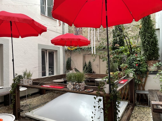 An outdoor patio area with several bright red umbrellas providing shade over wooden seating and tables. The space is surrounded by white walls and decorated with various potted plants, including small trees and flowering bushes. The floor is covered with small pebbles, and a skylight is integrated into the ground.