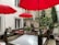 An outdoor patio area with several bright red umbrellas providing shade over wooden seating and tables. The space is surrounded by white walls and decorated with various potted plants, including small trees and flowering bushes. The floor is covered with small pebbles, and a skylight is integrated into the ground.
