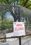 A chain-link fence with a prominent 'Area Closed' sign in front of lush green trees under a clear blue sky. The background reveals a portion of a significant white dome structure resembling a government building. The area appears to be secured as per the authority mentioned on the sign.