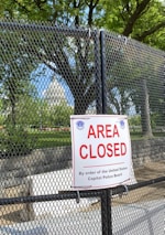 A chain-link fence with a prominent 'Area Closed' sign in front of lush green trees under a clear blue sky. The background reveals a portion of a significant white dome structure resembling a government building. The area appears to be secured as per the authority mentioned on the sign.
