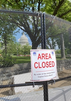 A chain-link fence with a prominent 'Area Closed' sign in front of lush green trees under a clear blue sky. The background reveals a portion of a significant white dome structure resembling a government building. The area appears to be secured as per the authority mentioned on the sign.