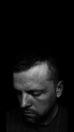 A black and white photograph featuring the upper half of a man's face, looking downward with eyes closed. The lighting creates a dramatic contrast, highlighting facial contours and texture of the skin while casting deep shadows.