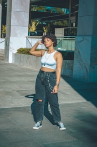 A person stands outdoors in an urban environment, wearing a white crop top and black, ripped jeans with a chain accessory. The background features modern architecture with glass and marble elements, along with some greenery. The sunlight casts long shadows on the pavement.
