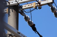 A close-up view of an electric utility pole with attached insulators and wires. The metal structure is mounted on a sturdy concrete pole, and the image captures the overhead power lines against a clear blue sky.
