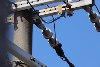 A close-up view of an electric utility pole with attached insulators and wires. The metal structure is mounted on a sturdy concrete pole, and the image captures the overhead power lines against a clear blue sky.