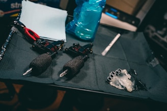 A set of tattoo equipment is neatly arranged, including two tattoo machines with black grips, a pair of red scissors, some wipes or paper towels, a blue spray bottle, and several small objects wrapped in plastic, possibly ink caps. The items are laid out on a dark, textured mat.