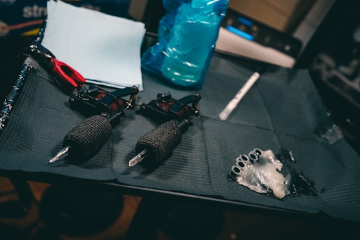 A set of tattoo equipment is neatly arranged, including two tattoo machines with black grips, a pair of red scissors, some wipes or paper towels, a blue spray bottle, and several small objects wrapped in plastic, possibly ink caps. The items are laid out on a dark, textured mat.